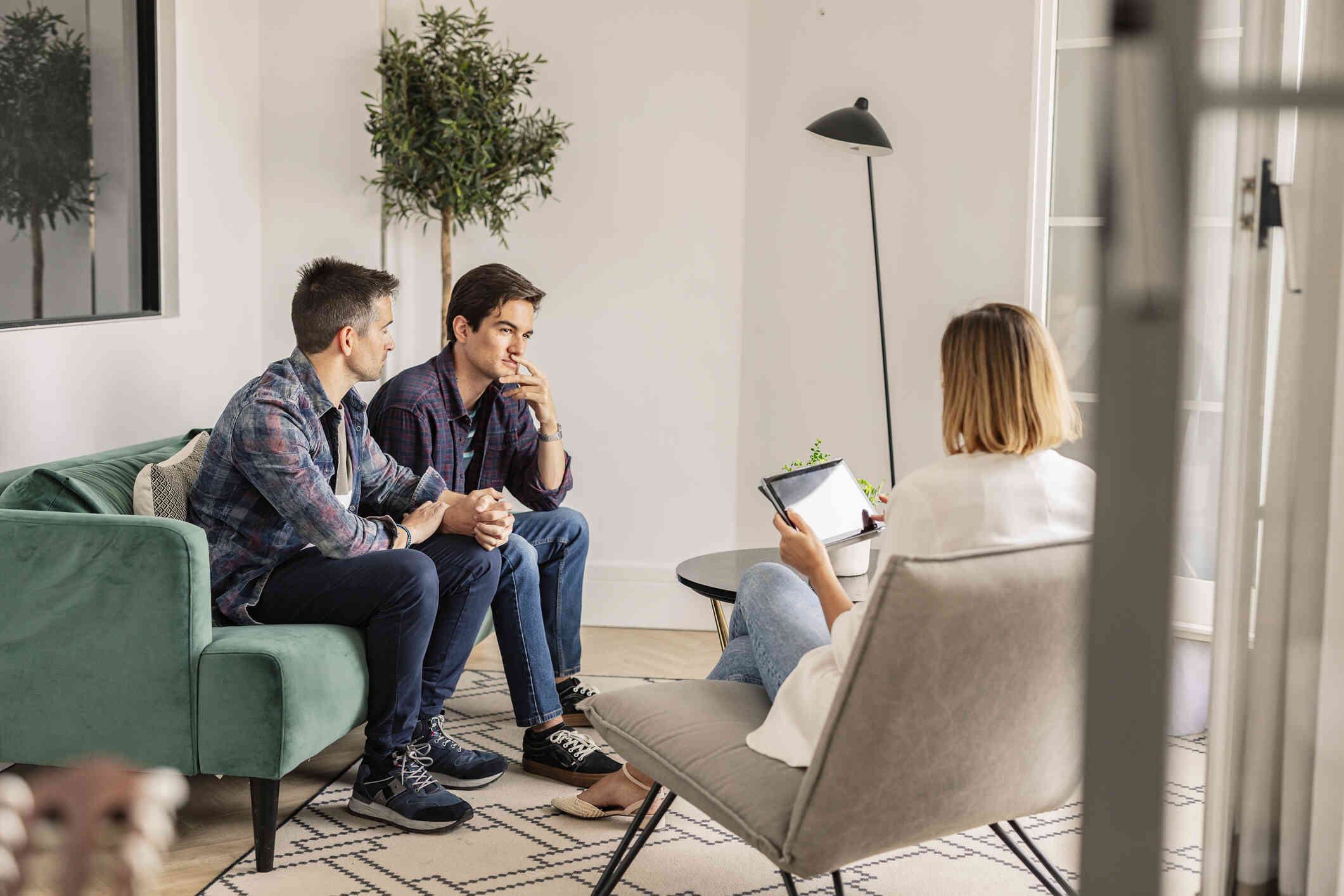 A couple sits together on a couch, attentively listening to a therapist holding a tablet.